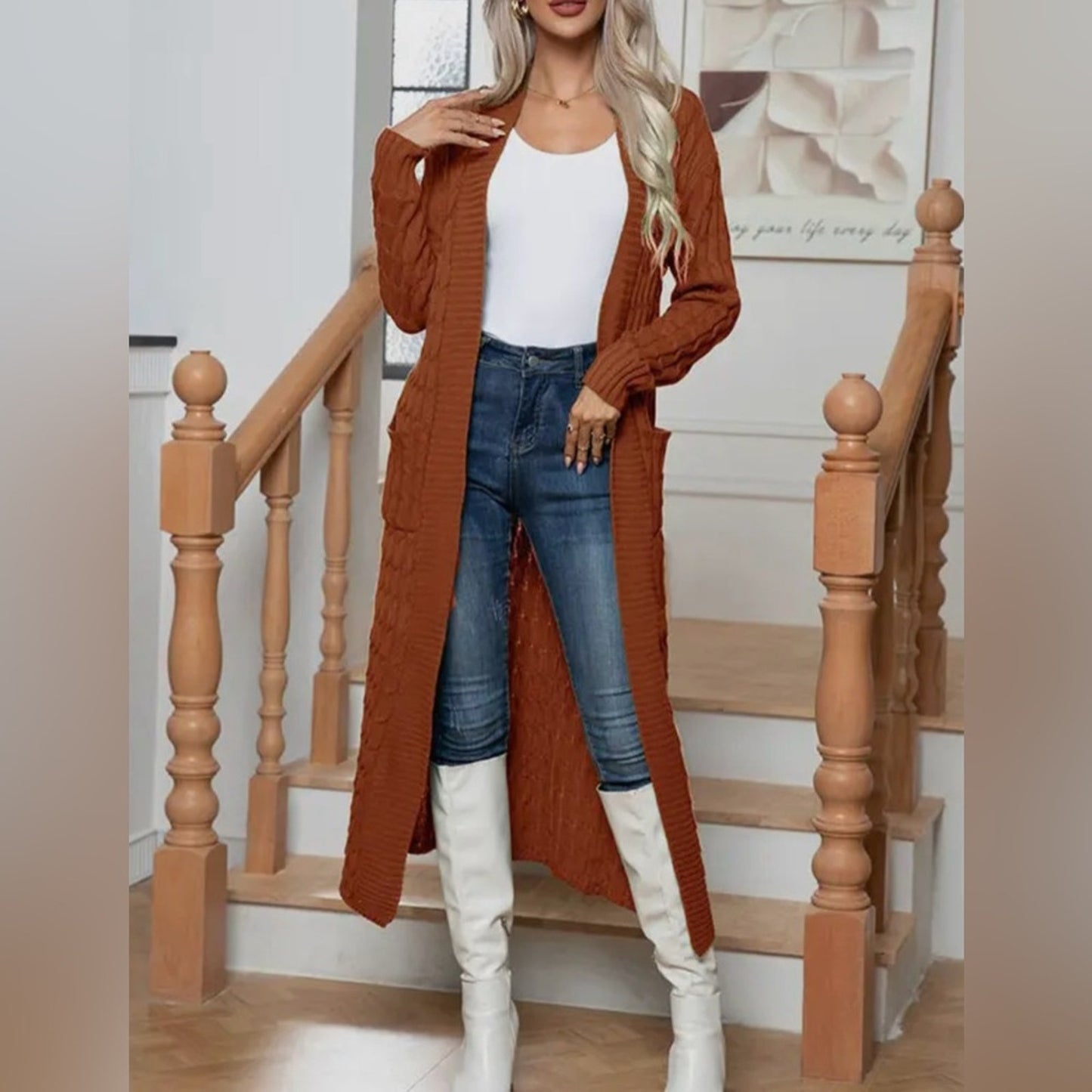 Woman wearing a long rust-colored cardigan, white top, blue jeans, and white knee-high boots standing in front of a wooden staircase.