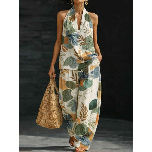 A woman models a sleeveless halter jumpsuit with a tropical leaf print in green, blue, and beige on an off-white base. She is holding a large woven straw tote bag against a dark studio background.