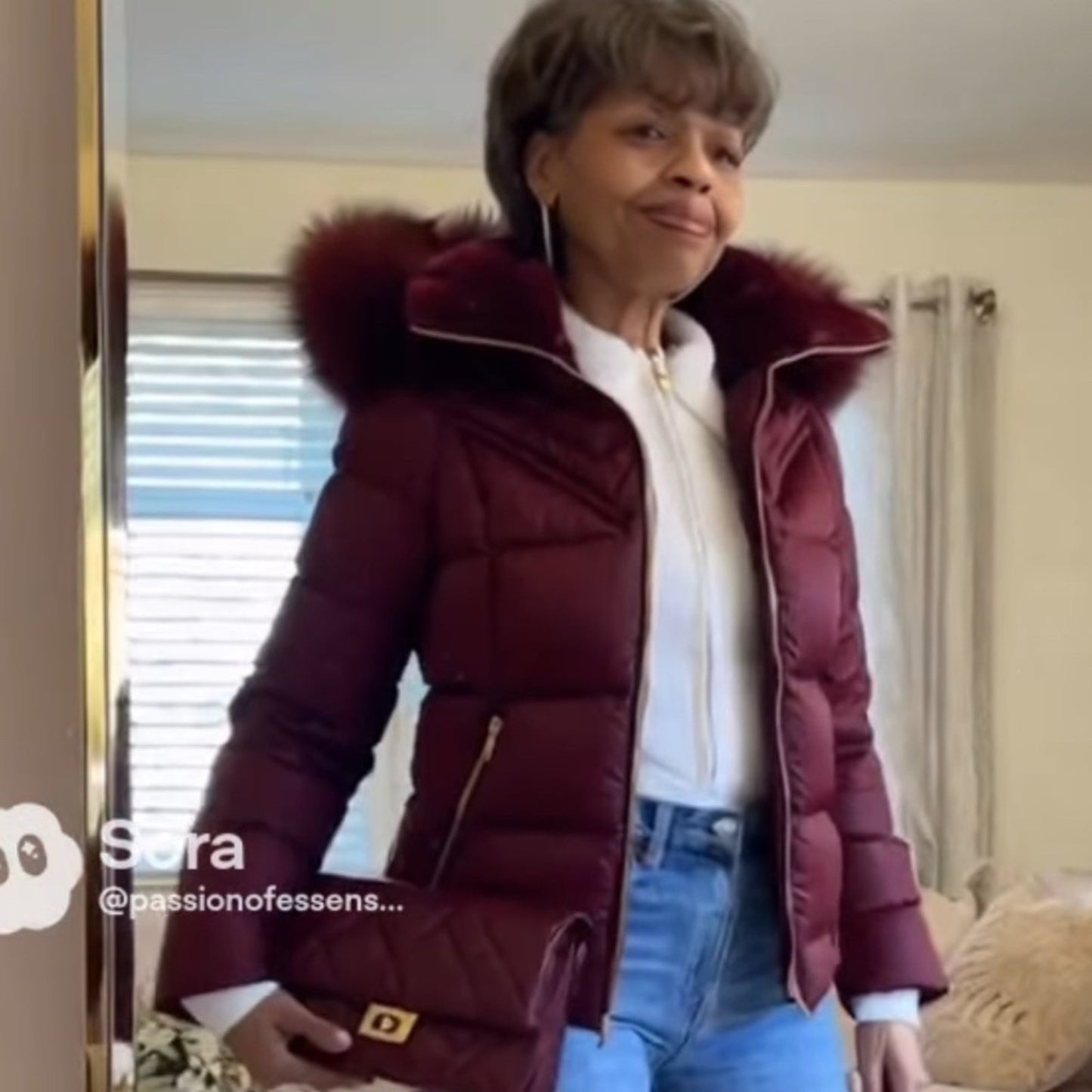 Woman wearing a maroon puffer jacket with a fur hood indoors.