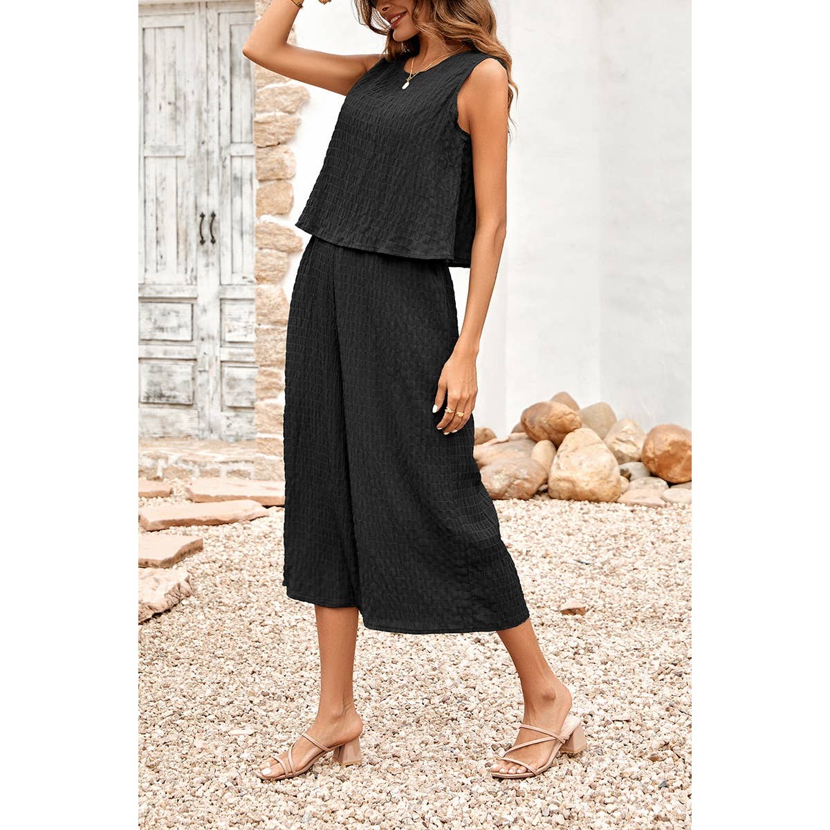 Woman wearing a black sleeveless top and wide-leg pants standing on pebbles with a stone wall background.