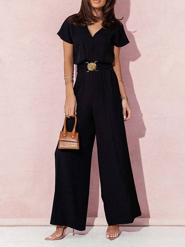 Woman wearing a black jumpsuit with a belt and holding a small brown handbag against a pink background