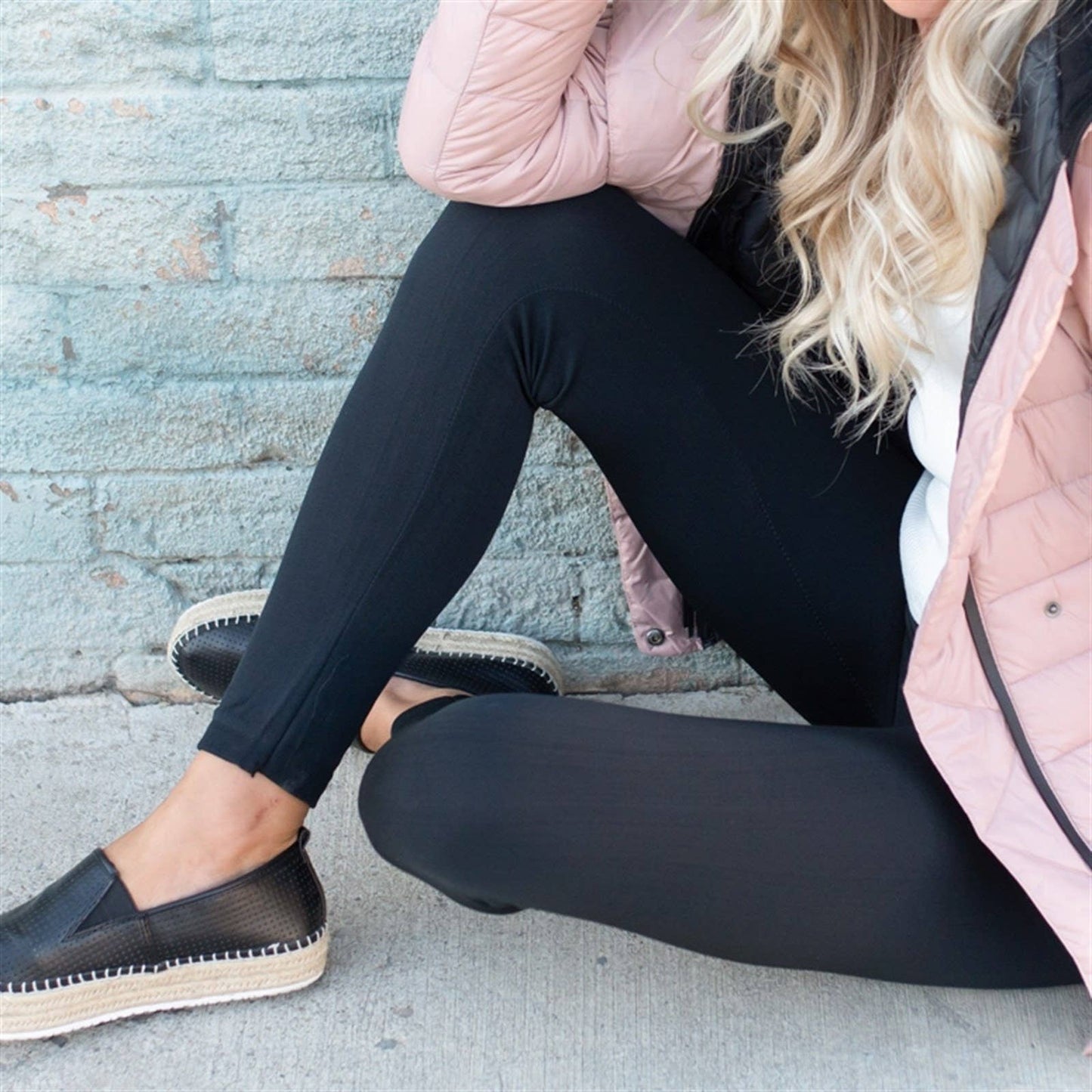 Person wearing black pants and a pink jacket sitting on a concrete surface.