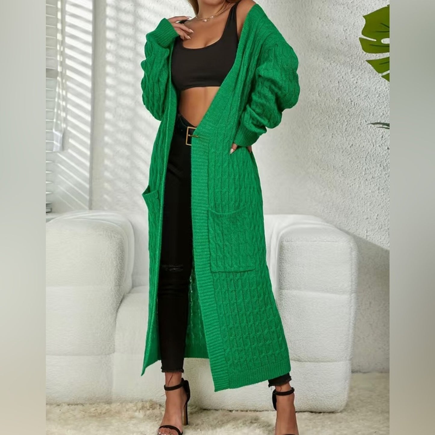 Person wearing a long green cardigan over black outfit in a minimal indoor setting