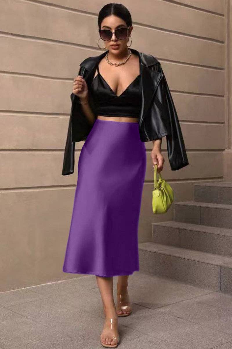 Woman in black leather jacket, black top, and purple skirt standing against a beige wall.