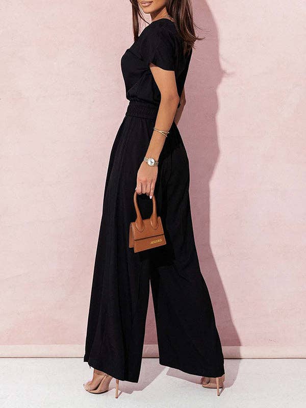 Woman wearing a black jumpsuit holding a brown handbag against a pink wall.