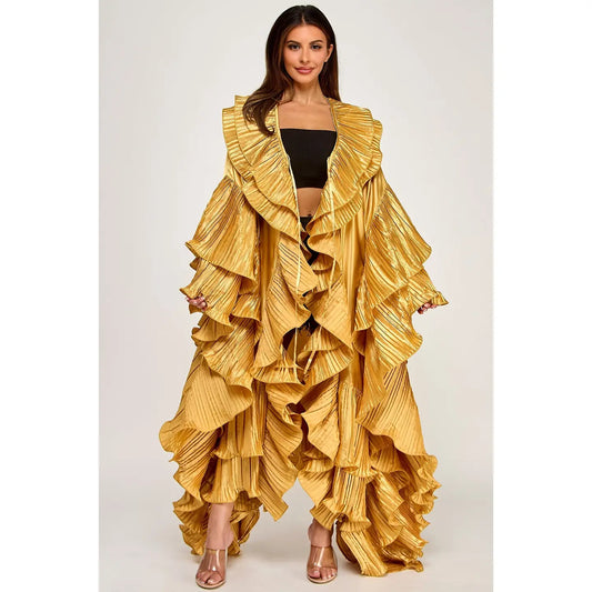 A model wearing a voluminous, metallic gold pleated ruffled jacket and matching pleated skirt, styled with a black crop top and metallic heels, against a light gray studio background.