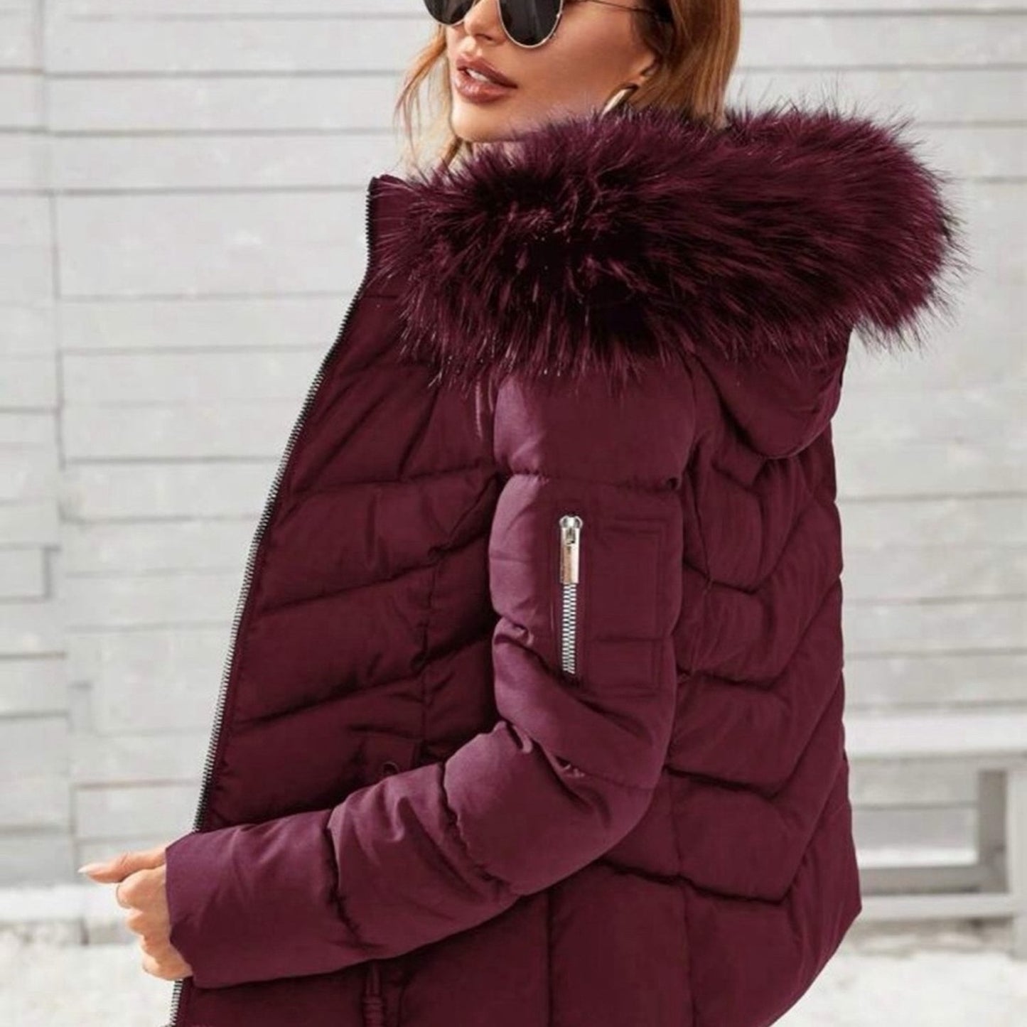 Person wearing a burgundy puffer jacket with a fur-lined hood against a light gray brick wall.