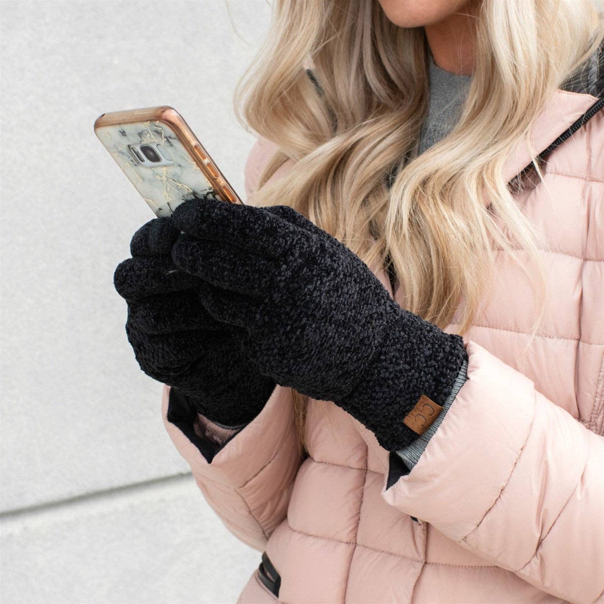 Person wearing black gloves holding a phone, with a light pink coat and blonde hair.