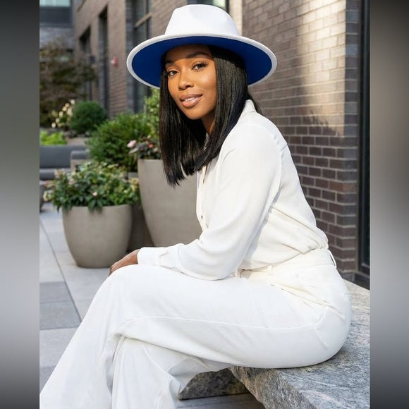 Woman in a white outfit and blue hat sitting outdoors near plants and a building.
