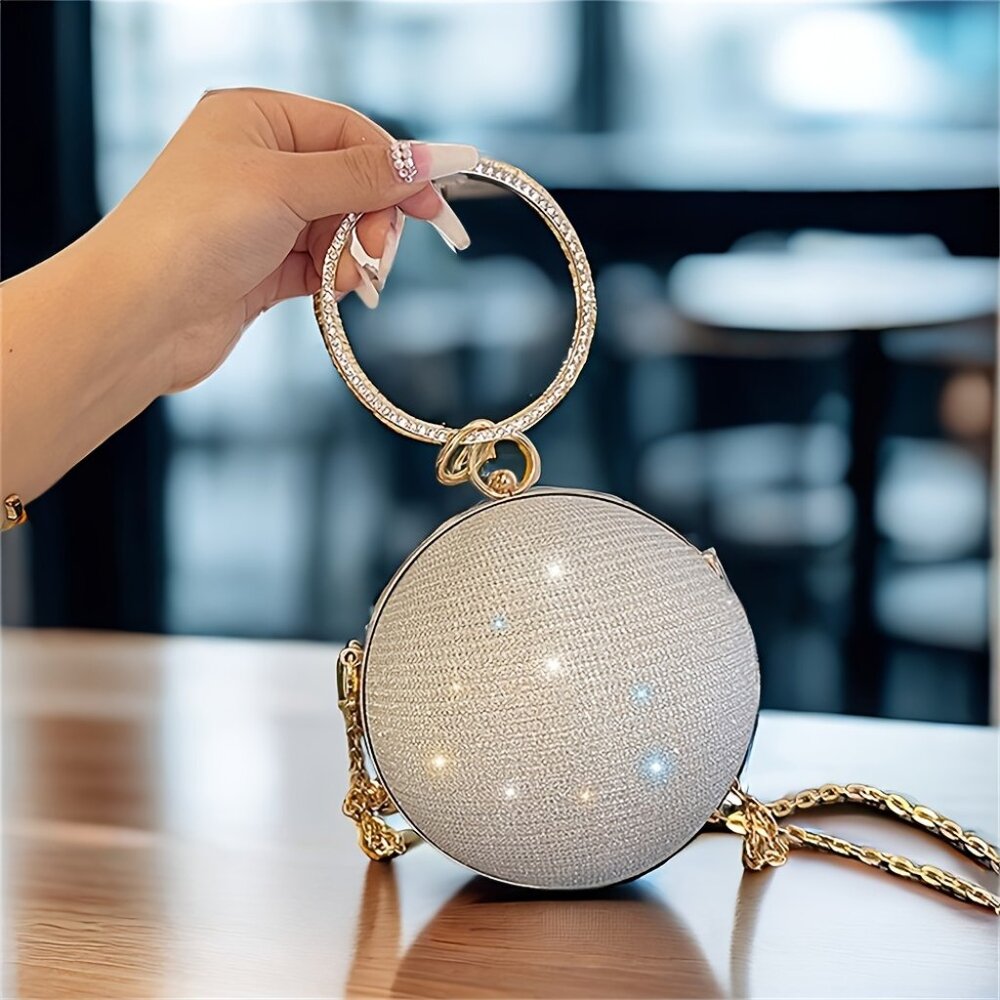 Hand holding a round, glittery handbag with a gold chain on a blurred indoor background